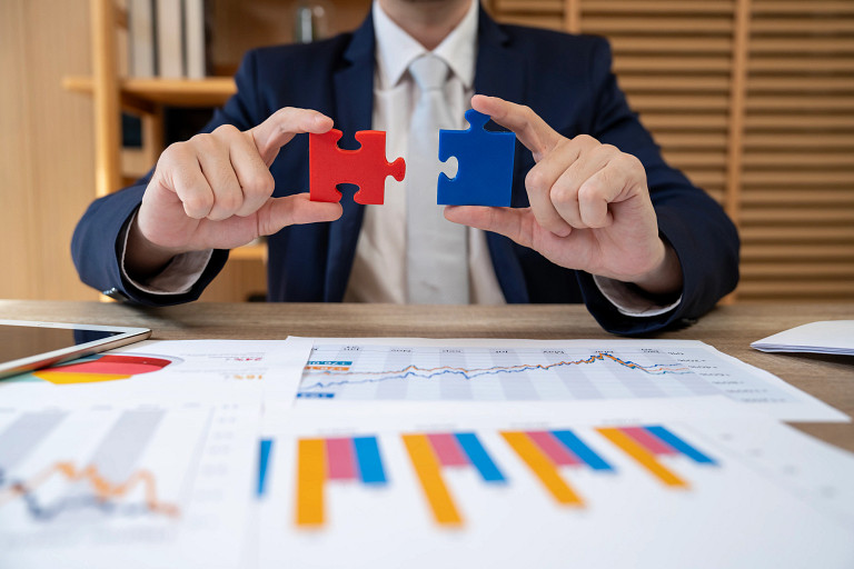 A business professional in a suit sitting at a desk holds two puzzle pieces, one red and one blue, symbolizing collaboration or strategy alignment. Financial charts and bar graphs are visible on the table, representing market analysis and investment planning.