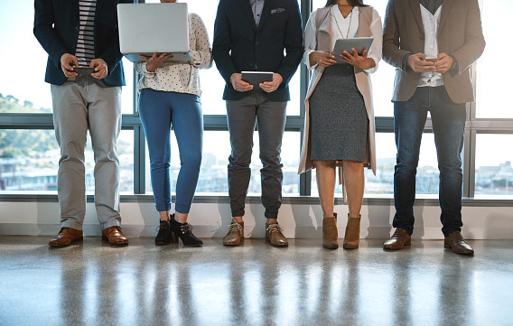 A group of five individuals standing indoors in front of large windows, shown from neck to feet. They are dressed in business casual attire and holding various devices, such as laptops, tablets, and smartphones. The polished floor reflects their footwear. The setting appears professional, possibly an office or conference space.