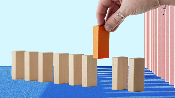 The image depicts a hand placing an orange block atop a line of beige wooden blocks. The blocks are arranged in a staggered formation, suggesting a domino effect.